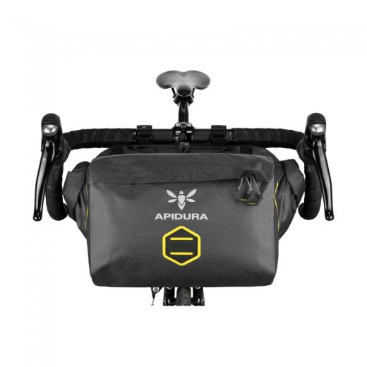 An Apidura Expedition Accessory Pocket 4.5L is shown in a grey waterproof laminate material, mounted between the drops of a gravel bicycle's handlebars