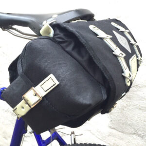 A Carradice Barley Seatbag is shown in a black canvas dick fabric with pale yellow straps and fitxtures, attached to the rails o a bicycle saddle
