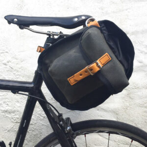 A Carradice Nelson Longflap Saddlebag is shown in a waxed black canvas material with leather trims hanging from the back of a saddle on a bicycle