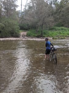A bikiepacker looks back over their shoulder at the camera as they push their bicycle across a wide but shallow river crossing to the gravel path in the distance
